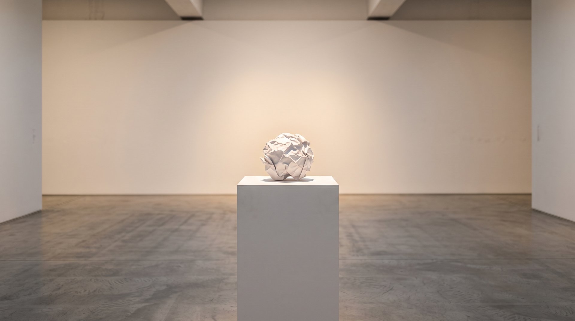 Una pallina di carta accartocciata esposta su un piedistallo bianco in una galleria d'arte vuota, sotto un faretto caldo